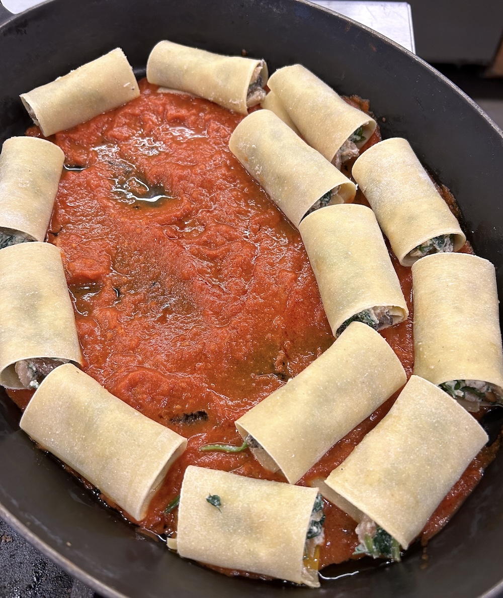Uncooked lasagna noodles, rolled up around filling, sit in a red sauce in a black dish